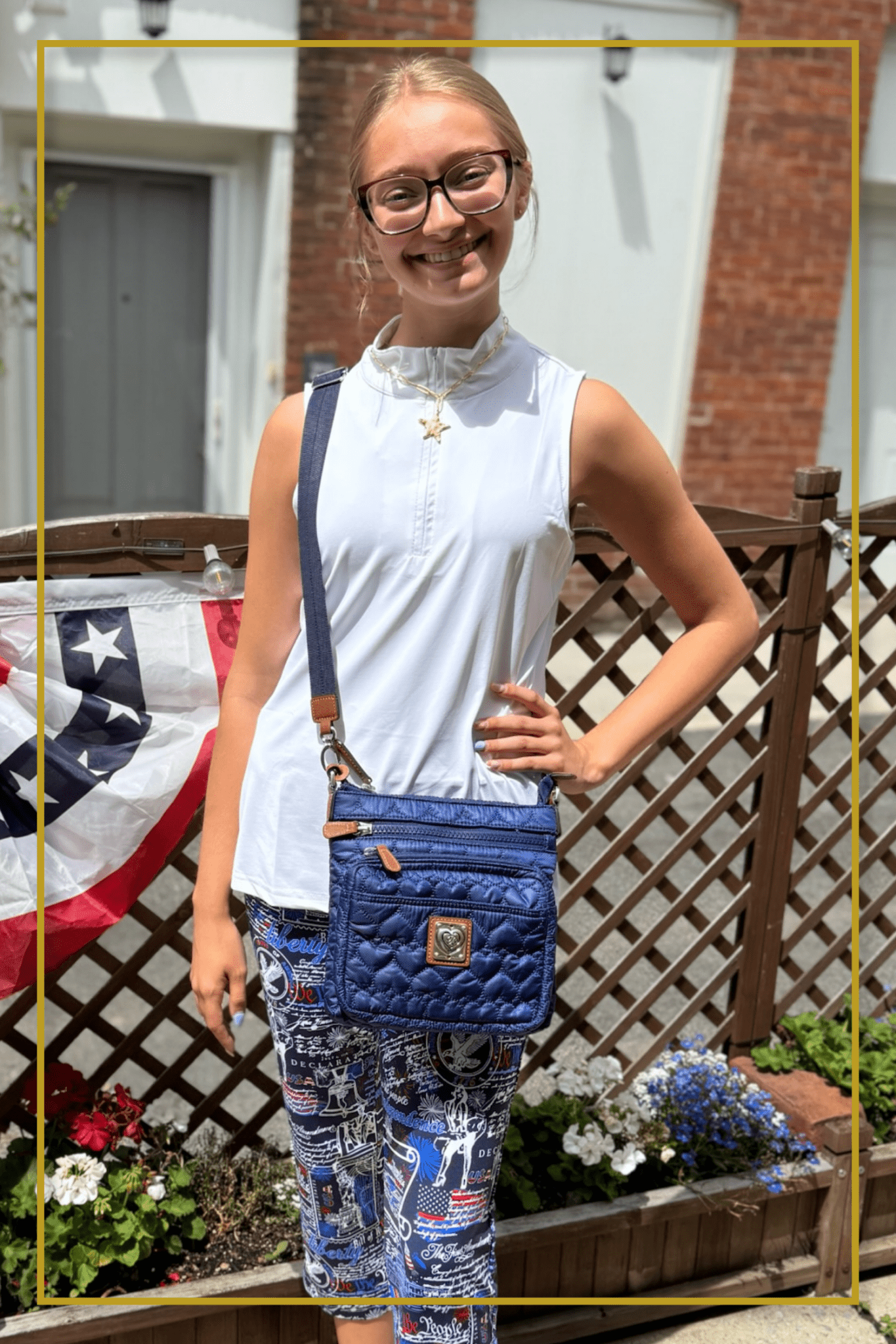 Woman in a white quarter-zip top and patriotic print leggings carrying a navy quilted crossbody bag near an American flag